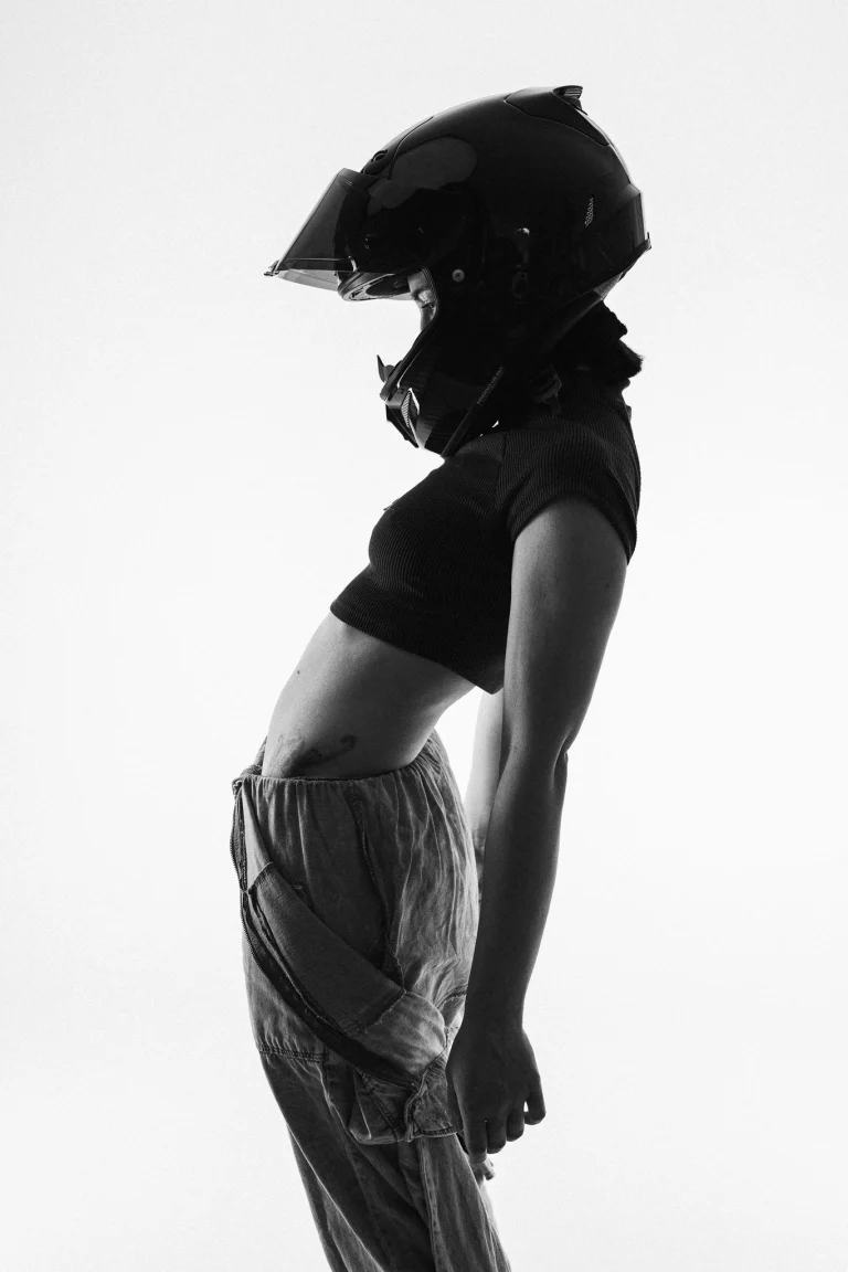 A woman wearing a black motorcycle helmet and a crop top arches her back against a bright white background in this black and white portrait.