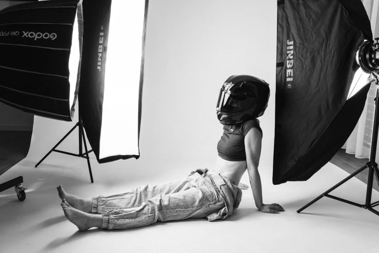 A woman wearing a black motorcycle helmet and loose trousers sits on the floor in a brightly lit studio between two large softboxes.