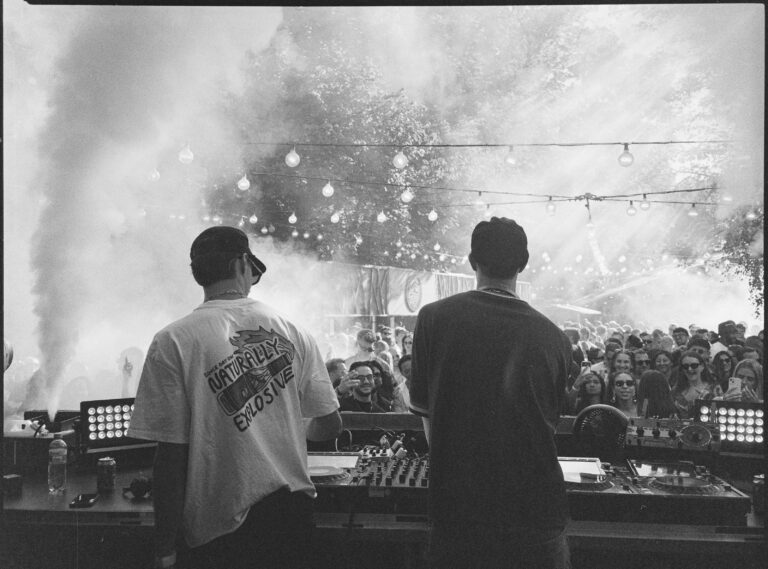 Monochrome, grainy analog photograph of two men DJing from behind the booth at an outdoor festival, with a thick plume of smoke rising on the left and a crowd visible in the background under string lights.