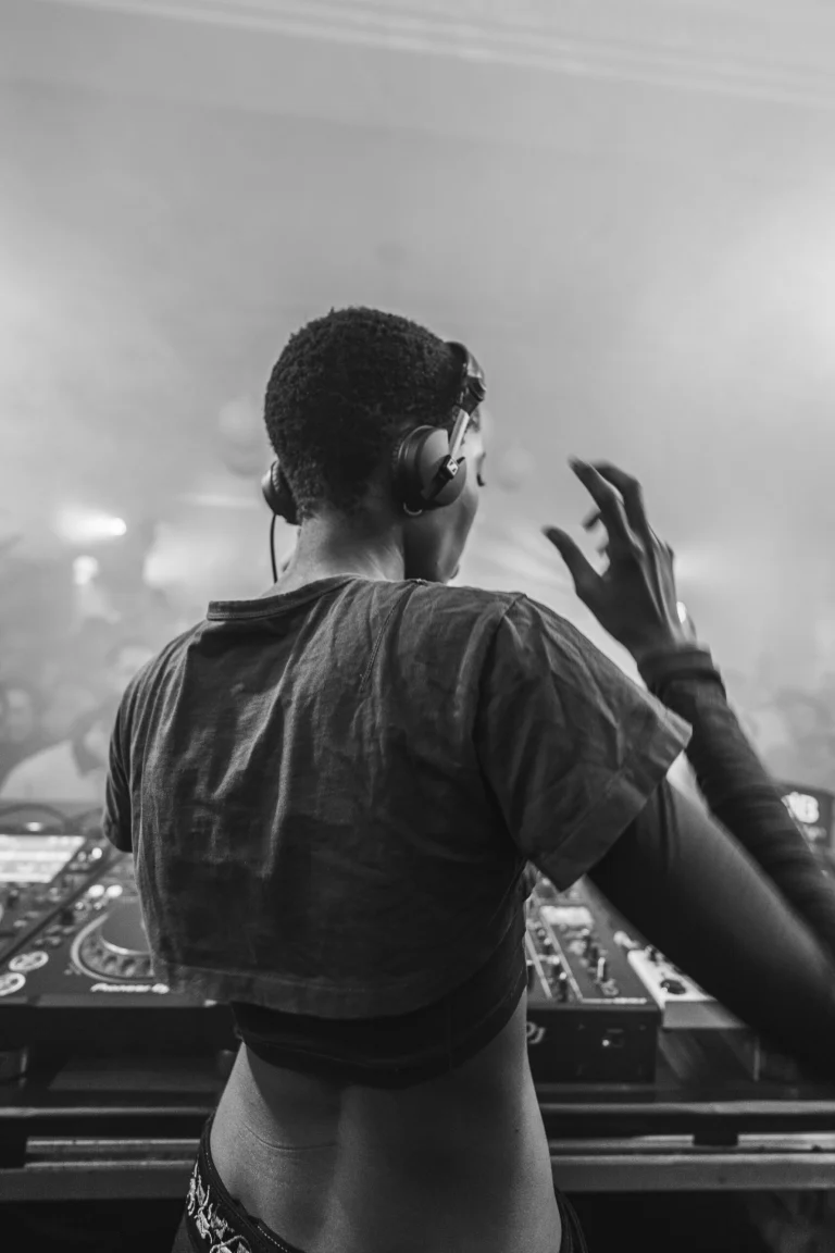 A woman with short hair wearing headphones stands with her back to the camera, gesturing over a DJ mixer in a monochrome club setting.