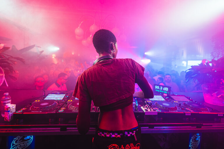 A woman stands behind DJ equipment, facing a crowd in a room filled with pink and blue stage lighting and smoke.