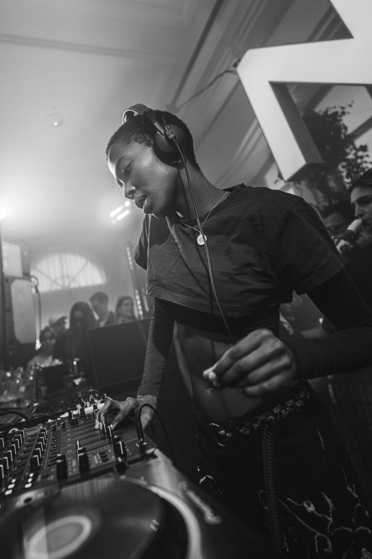 A woman wearing headphones and dark clothing stands behind a mixing console, adjusting knobs in a brightly lit indoor venue.