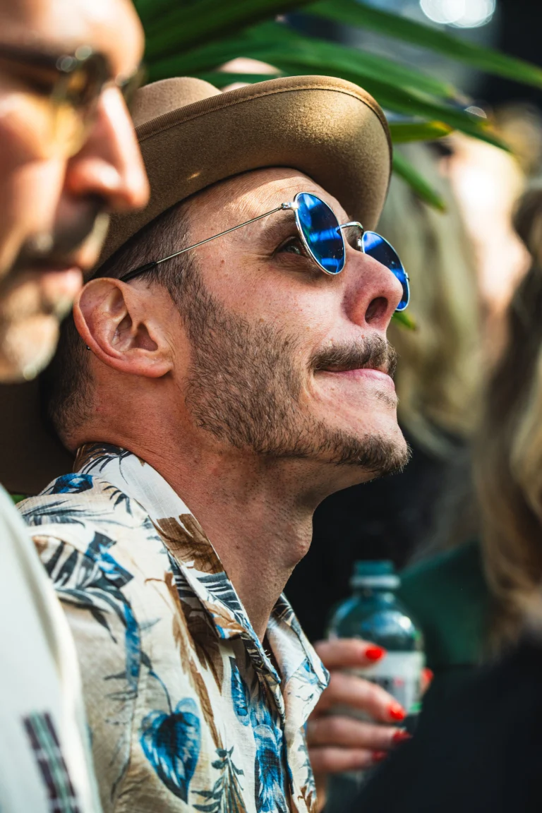 A man wearing a hat, blue-tinted sunglasses, and a patterned shirt looks upward in natural light.