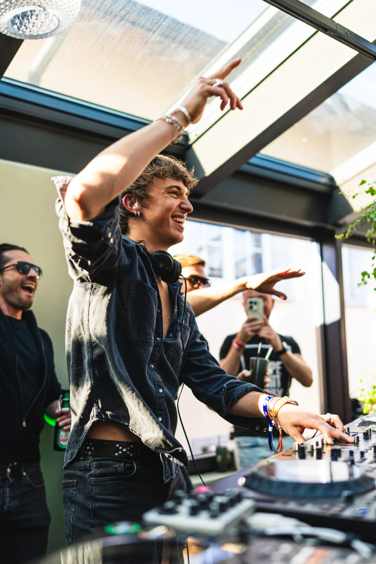 A smiling man stands behind a DJ mixer indoors, raising his arms beneath a glass ceiling while other people gather in the background.