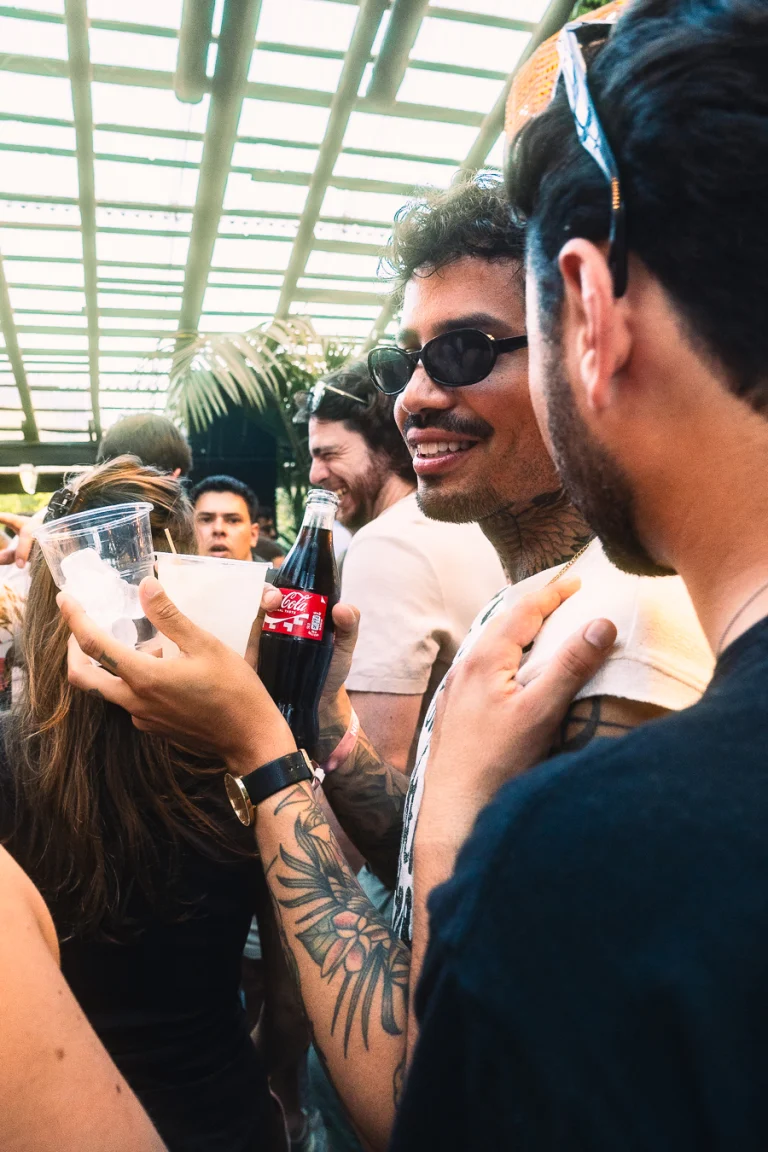 A man holds drinks and a glass bottle of Coca-Cola outdoors while other men stand nearby under a slatted roof.