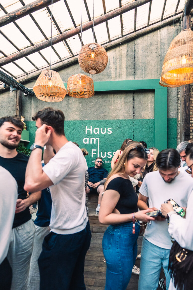 Several men and women gather under woven pendant lamps in a rustic hall with a green wall bearing the text Haus am Fluss.