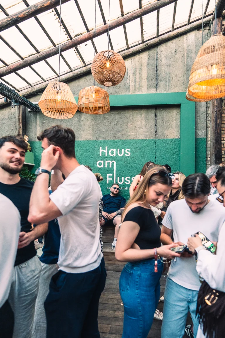 Several men and women gather under woven pendant lamps in a rustic hall with a green wall bearing the text Haus am Fluss.