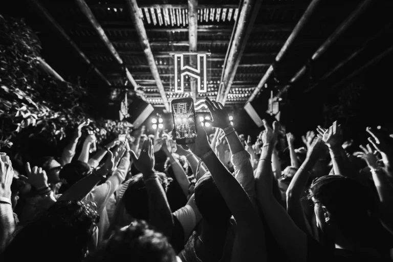 A black-and-white shot of a crowd at Haus am Fluss, with a person holding up a phone to record and hands raised in the dim venue.