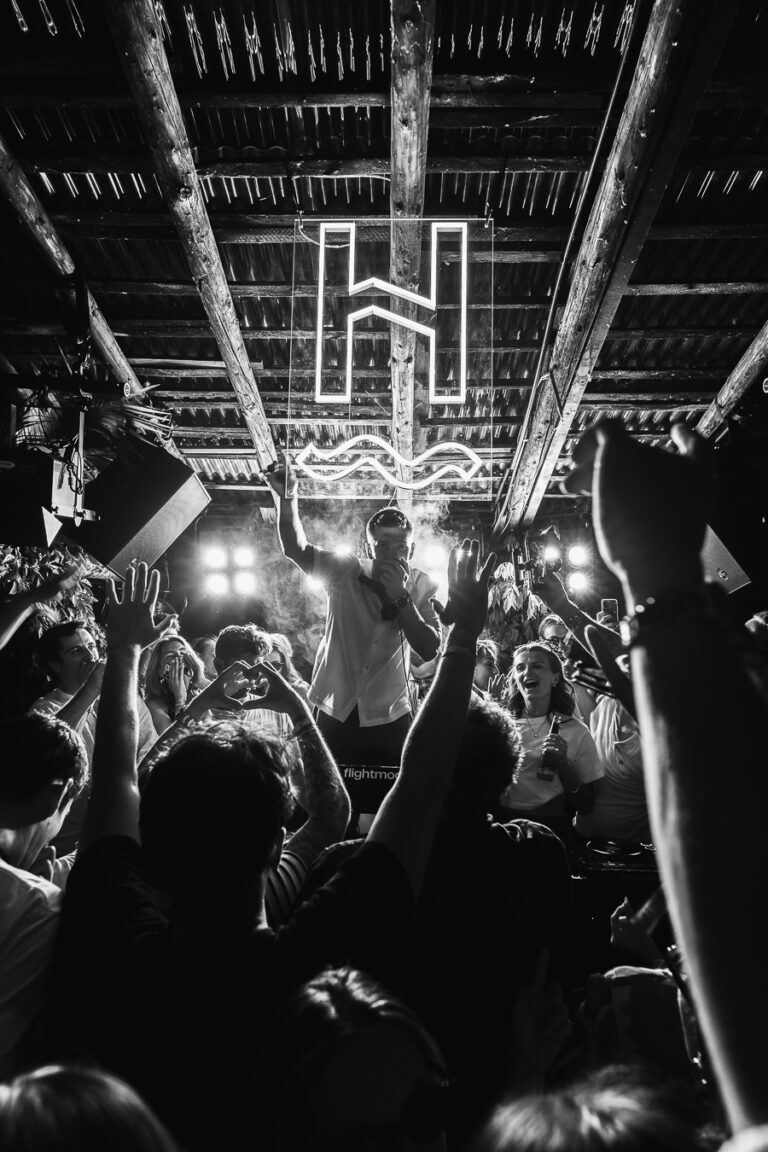 A black-and-white shot of a man performing on stage beneath a glowing neon sign, surrounded by a raised-hands crowd.