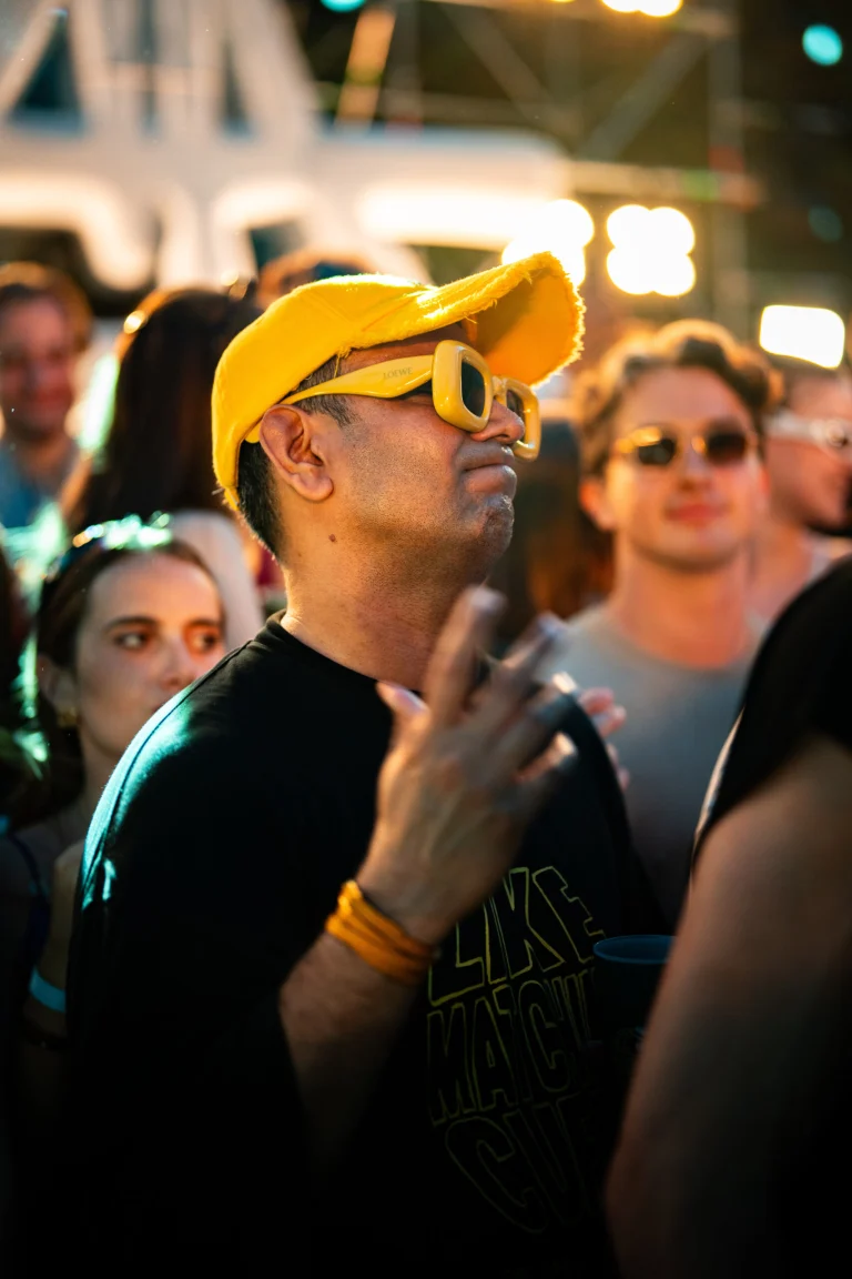 A man in a yellow cap and sunglasses gestures amidst a crowd at an outdoor night event.