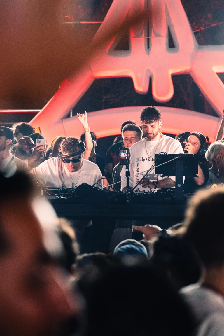 Two men stand behind a DJ mixing console under red stage lighting, with a crowd visible in the foreground and background.