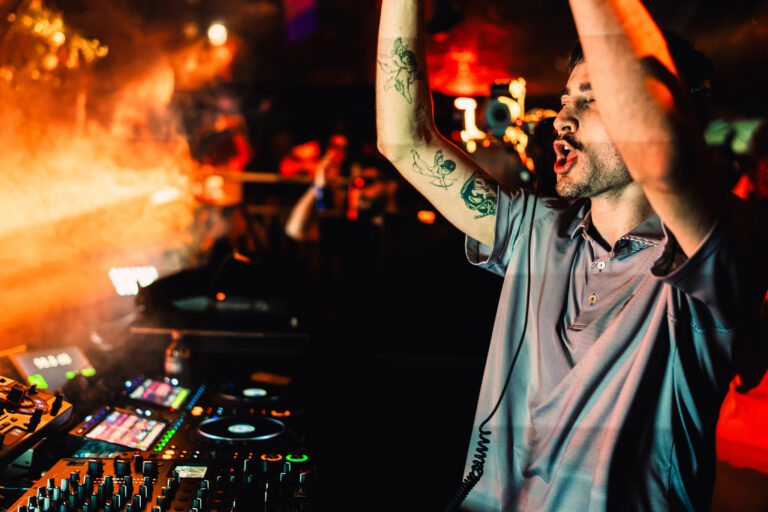 A man with tattoos on his arm stands behind a DJ mixer with his hands raised, under warm stage lighting in a dimly lit club.