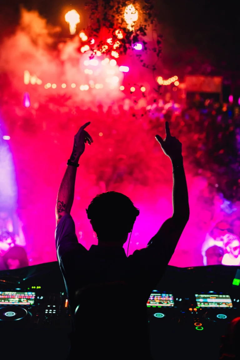 A man stands behind a mixing console with his hands raised in front of a brightly lit stage filled with red smoke and a crowd.