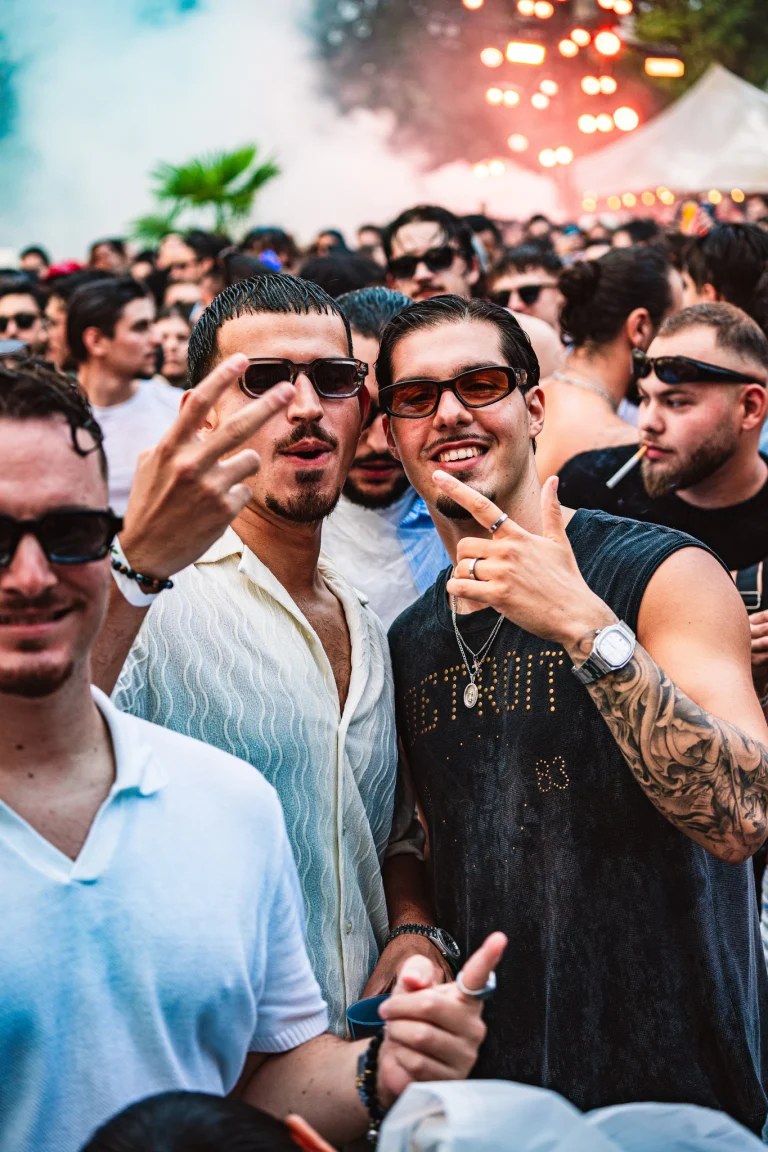 Three young men pose closely together outdoors in a dense crowd at a festival, flashing hand signs and wearing sunglasses.