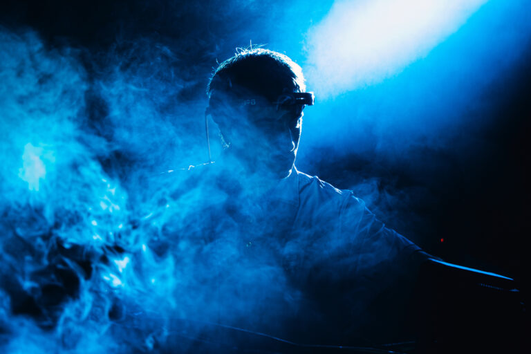 A man stands in thick blue stage smoke illuminated by a bright overhead spotlight.