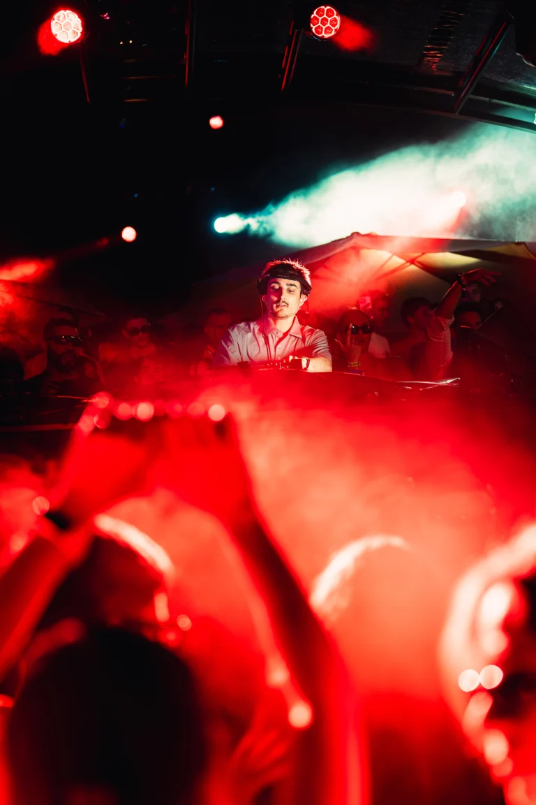 A man stands behind a DJ booth in a dark venue filled with red smoke and stage lighting, with silhouetted hands raised in the foreground.