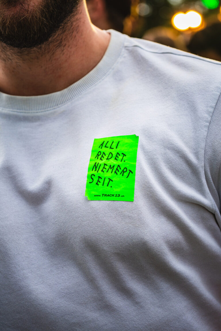 A man wearing a white t-shirt with a bright green sticker on the chest featuring handwritten text reading ALLI REDET. NIEMERT SEIT.