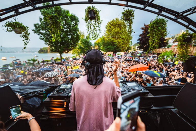 A man wearing a pink shirt and a black head covering stands behind a mixing console, facing a large crowd gathered outdoors by a lake.
