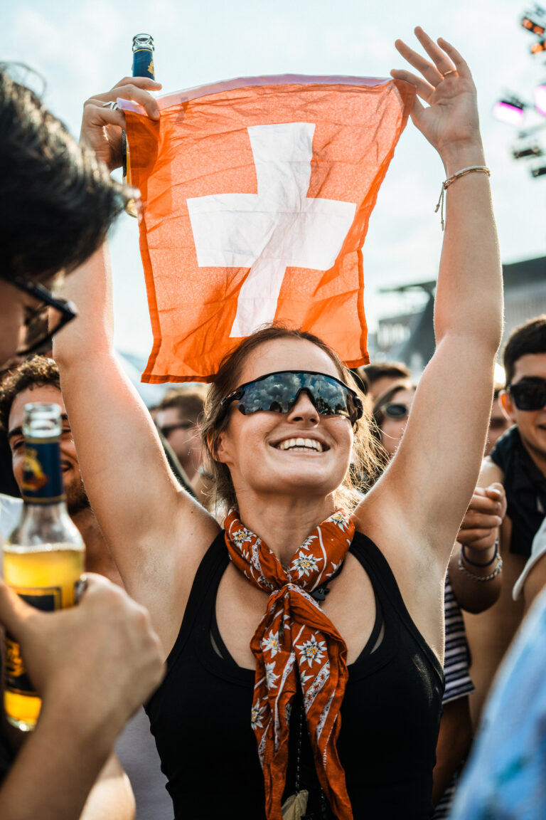 A woman wearing sunglasses holds up a Swiss flag above her head in an outdoor crowd.