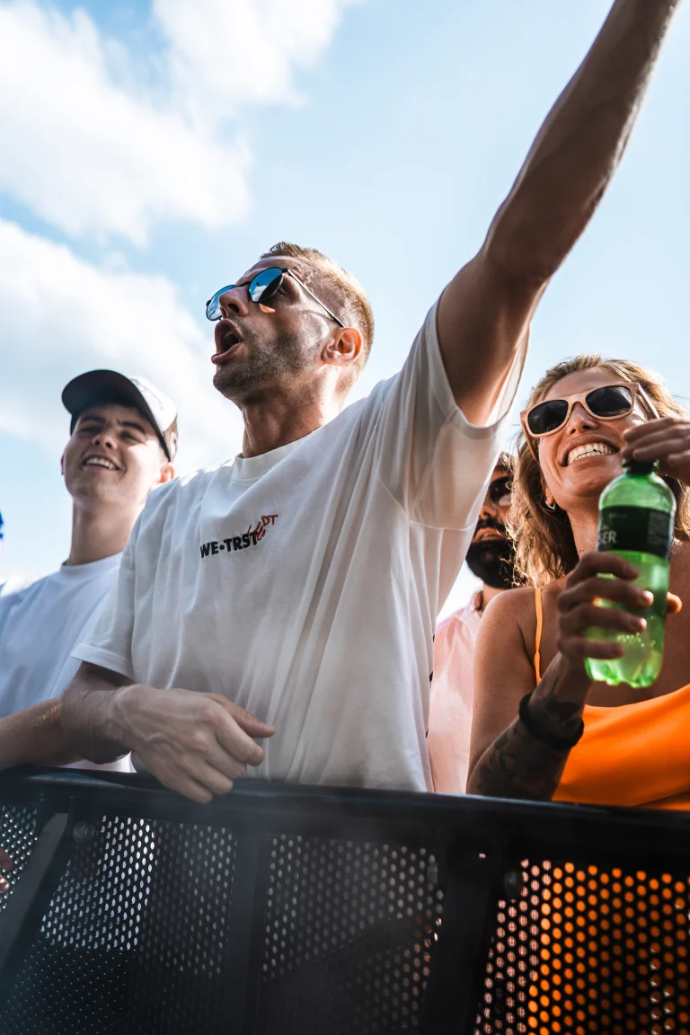 A man in a white t-shirt and sunglasses raises his arm in front of a metal barrier under bright daylight, with a woman holding a green bottle beside him.