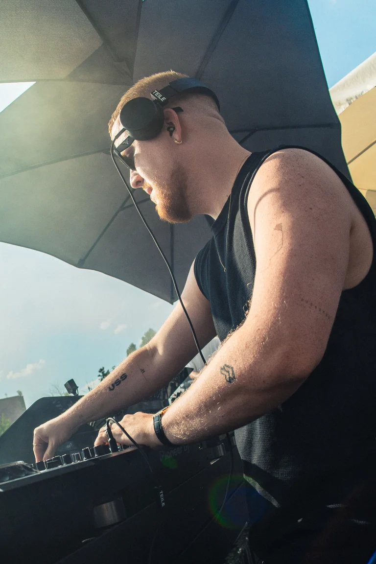 A man wearing headphones operates a mixing console under a large umbrella in daylight.
