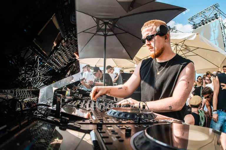 A man wearing sunglasses and headphones operates a DJ mixing console outdoors under a sun umbrella, with a crowd visible in the background.