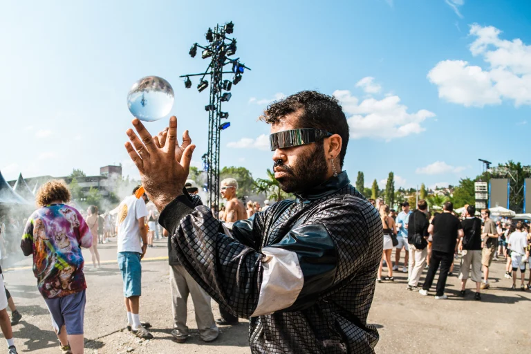 A man wearing futuristic mirrored visors balances a glass sphere above his open hands outdoors in natural daylight, with a crowd and stage equipment in the background.