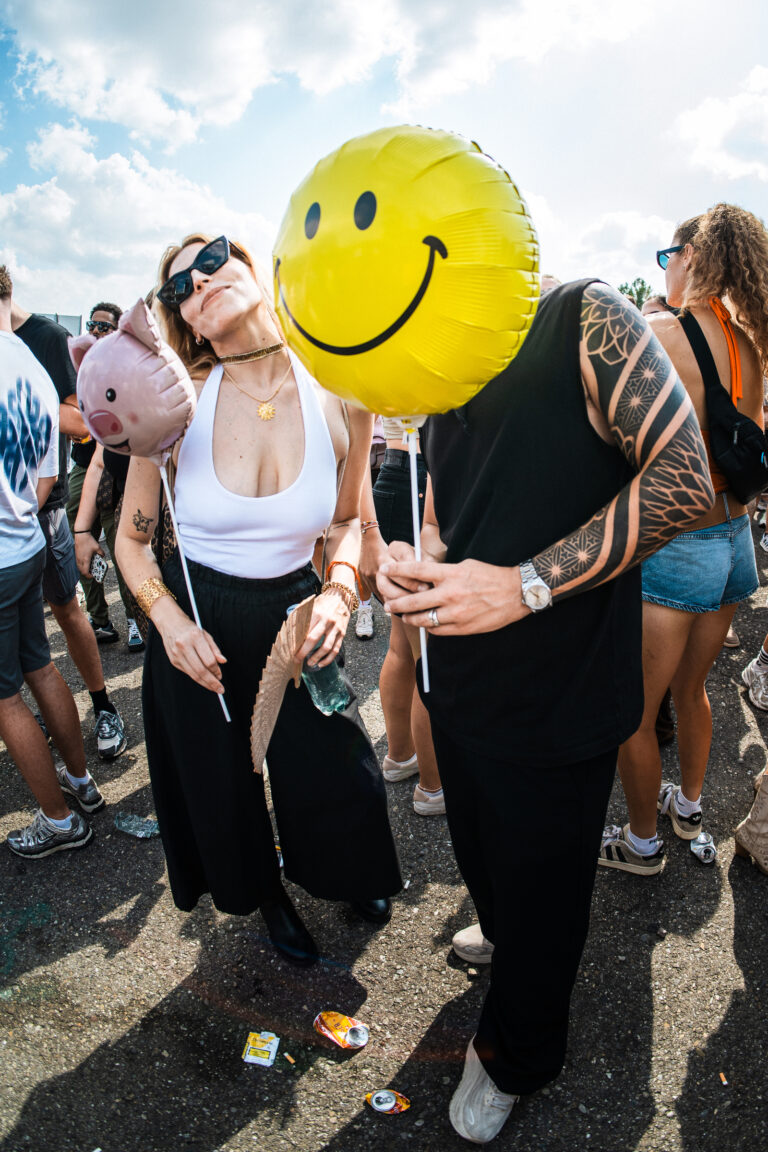 A woman wearing sunglasses and a white top holds a small pig-shaped balloon, while a man next to her holds a large yellow smiley face balloon.