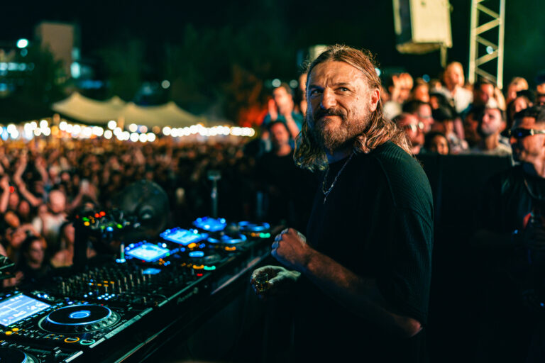 A long-haired man with a beard stands behind a DJ mixing console at night, looking back toward a crowded outdoor festival audience.