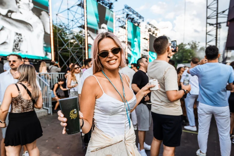 A smiling woman wearing sunglasses and a white top holds a cup in a crowded outdoor festival setting.