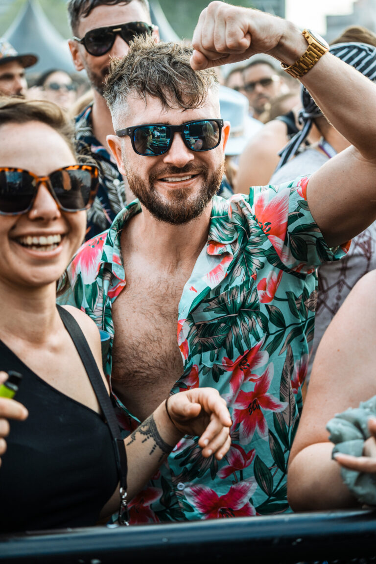 A smiling man with sunglasses and a floral shirt raises his fist in a crowd, with a smiling woman in the foreground.