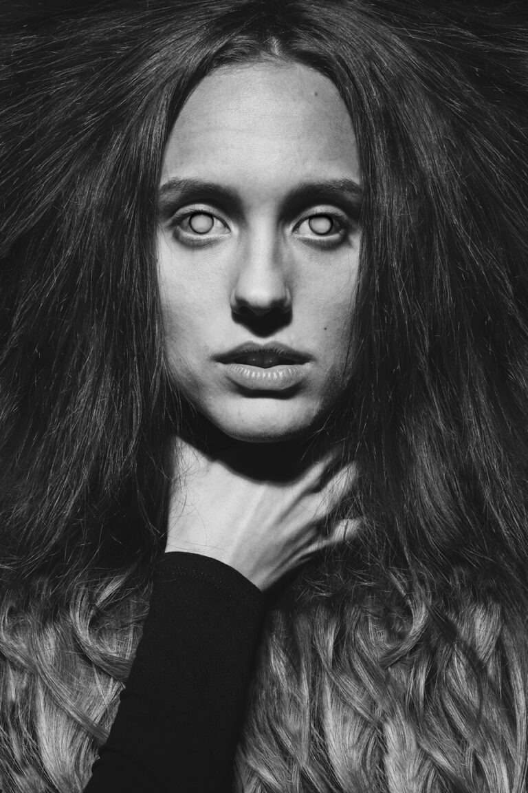 Close-up black and white portrait of a woman with white glowing eyes and dark hair fanned out, holding her own neck with one hand.