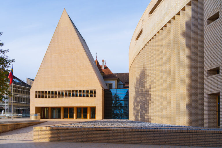 Exterior view of the Vaduz Parliament building showing a triangular brick structure connected to a curved wing by a glass facade.