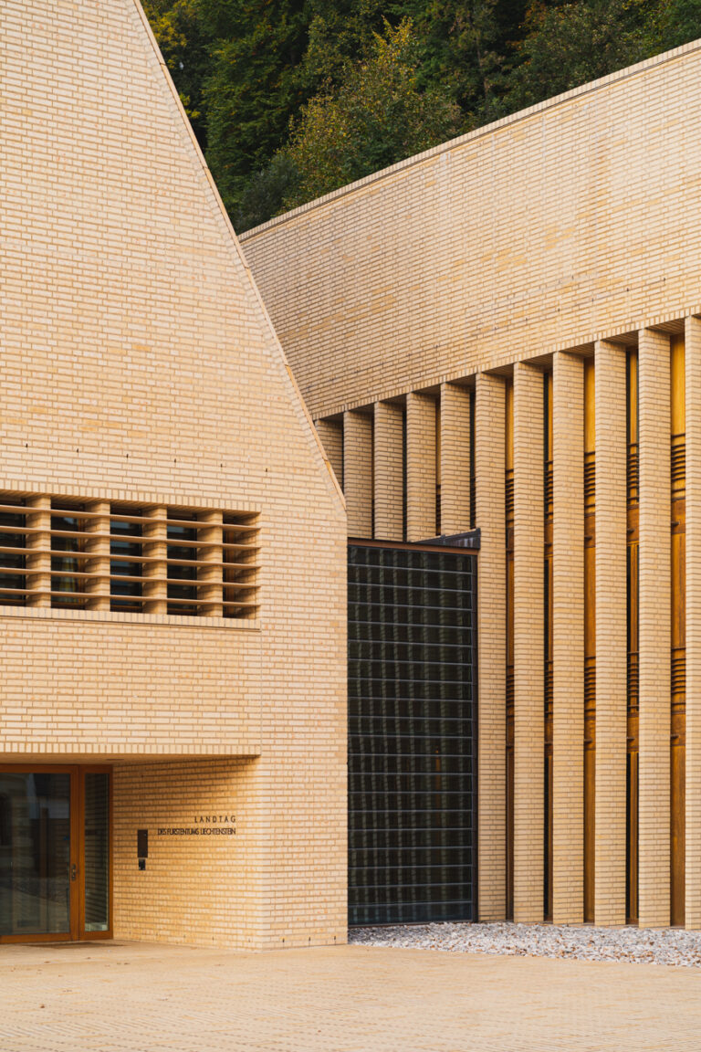 Light brick facade of the Vaduz Parliament building with sharp geometric angles, tall vertical columns, and a dark gridded window section.