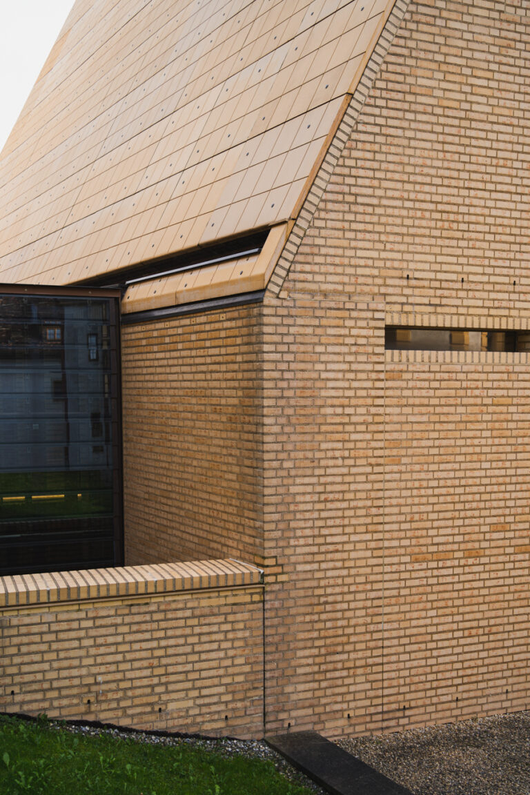 Close-up of a modern brick building with a steep tiled roof and a glass facade reflecting other buildings.