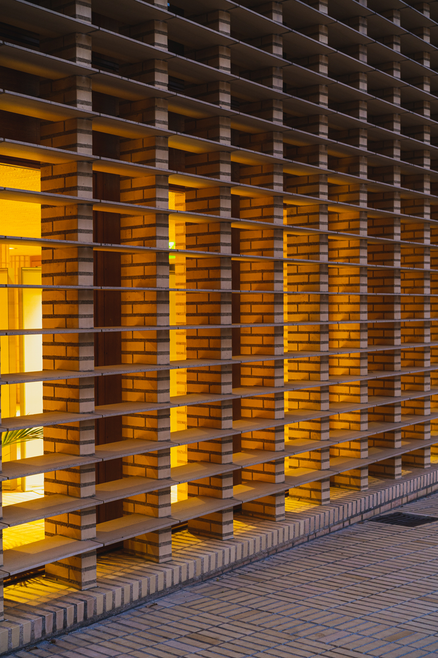 Eine detailreiche Fassade zeigt ein geometrisches Raster aus gestapelten Ziegeln und horizontalen Lamellen, wobei warmes Licht aus den Innenfenstern scheint.