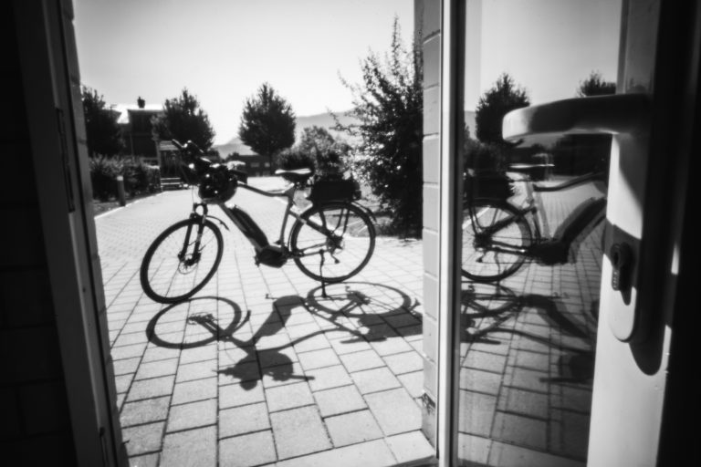 Schwarzweiß-Weitwinkelaufnahme eines Fahrrades auf einem gefliesten Innenhof, gespiegelt in einer Glastür mit einem auffälligen Griff.