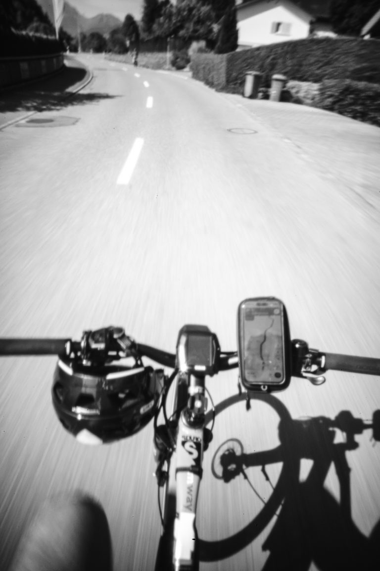 Monochrome Ich-Perspektive vom Fahrradlenker aus bei der Fahrt auf einer kurvigen Asphaltstraße mit Bewegungsunschärfe.