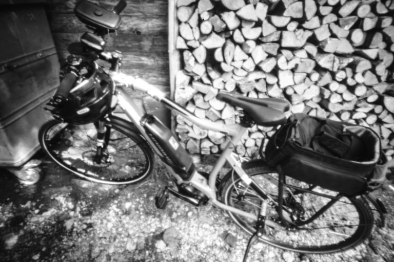 Monochrome, leicht verzerrte Analogaufnahme eines Fahrrads, das vor einem gestapelten Holzhaufen geparkt ist.