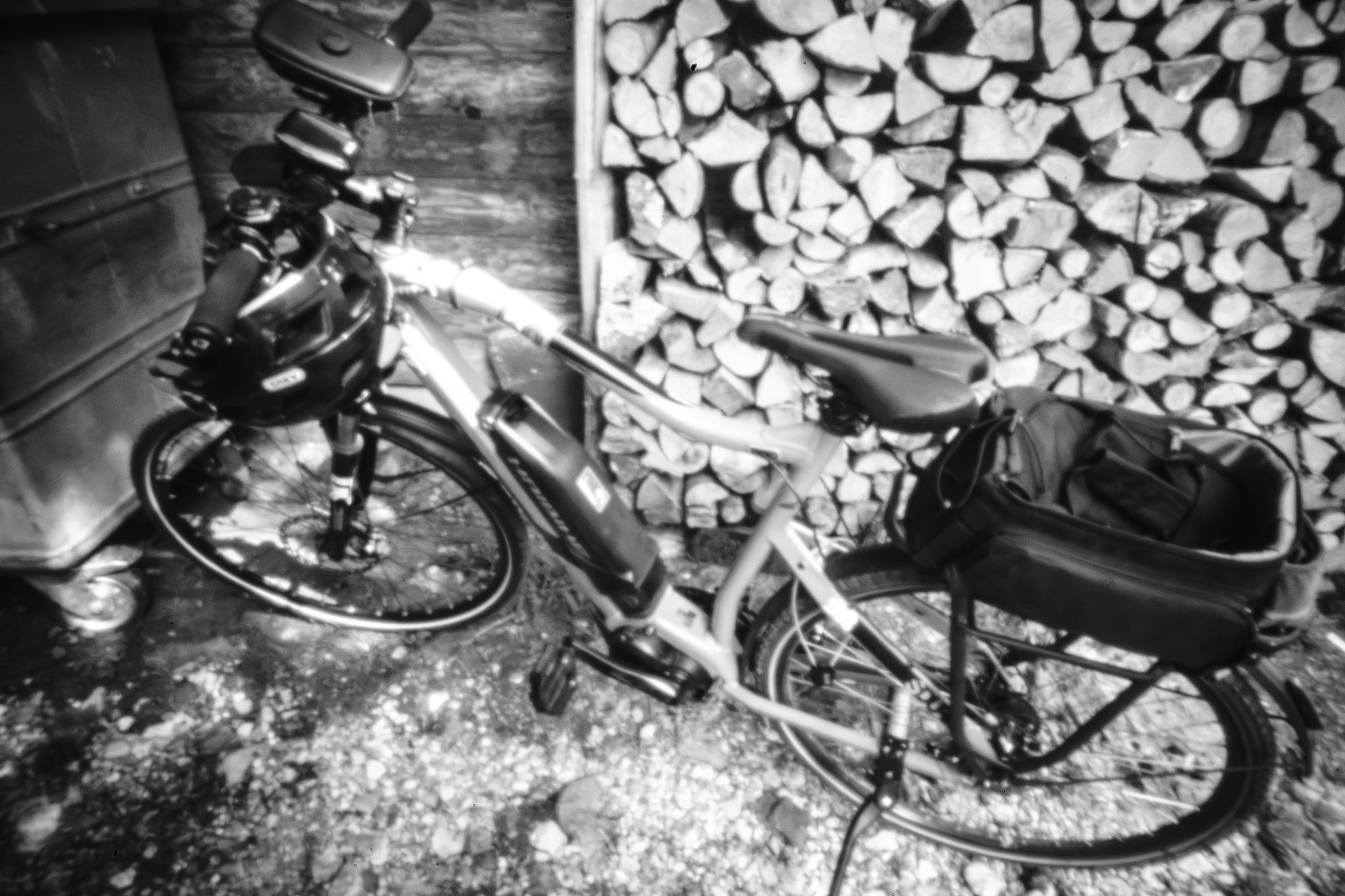 Monochrome, leicht verzerrte Analogaufnahme eines Fahrrads, das vor einem gestapelten Holzhaufen geparkt ist.
