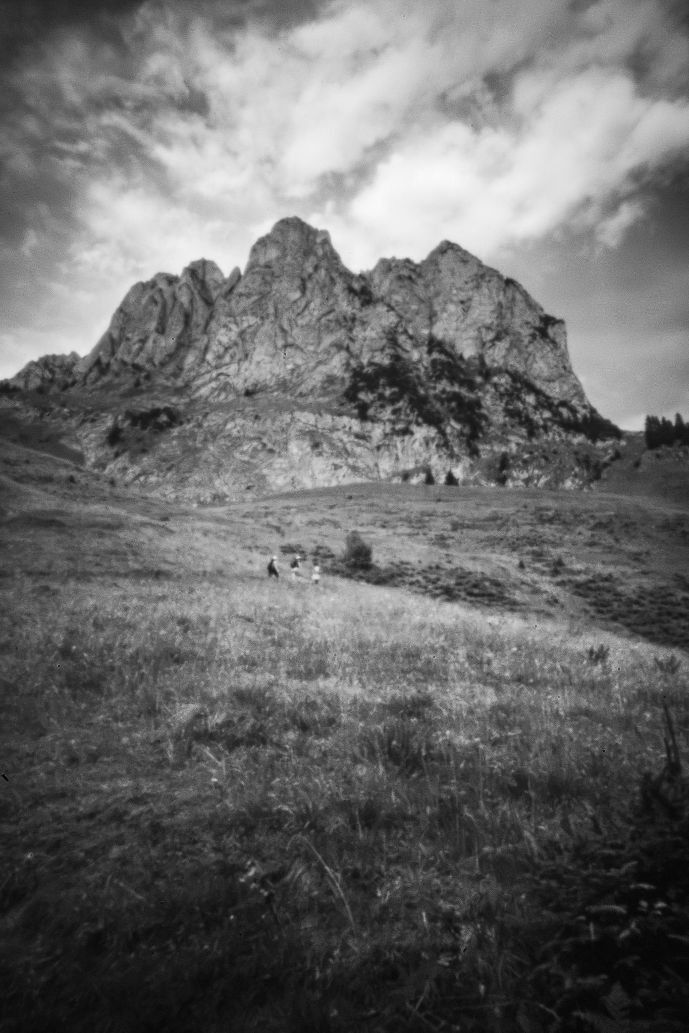 Monochrome Lochkamera-Aufnahme von gezackten Berggipfeln über einem Grasanghang mit drei winzigen Gestalten in der Ferne.