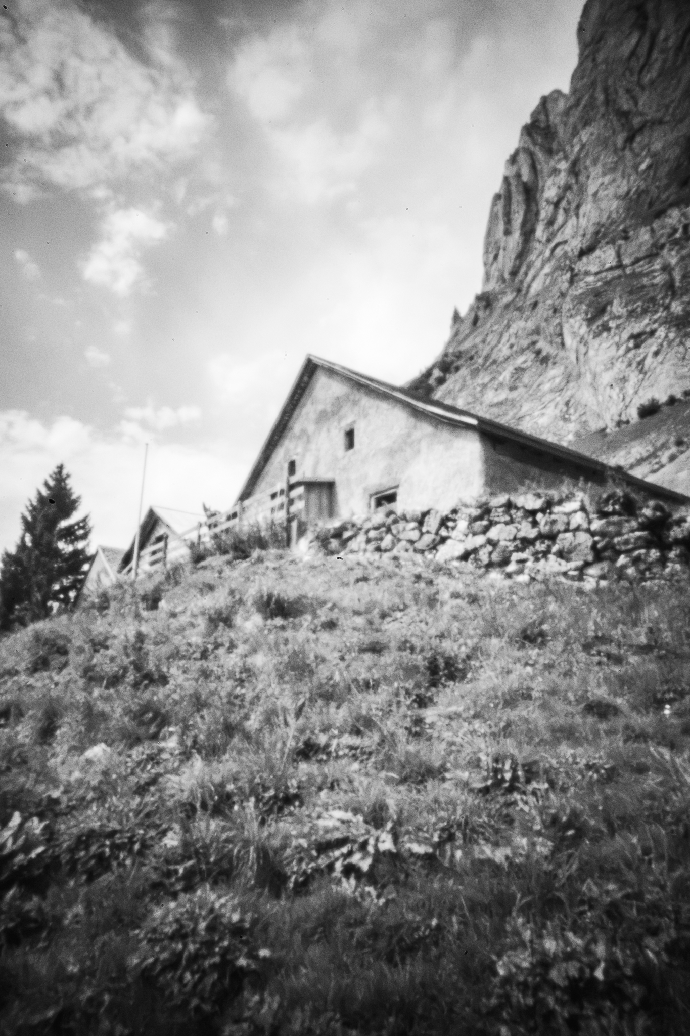 Monochrome Lochkameraufnahme von alpinen Steinhäusern auf einem grasbewachsenen Hang unter einer Felswand, mit unscharfem Rand.