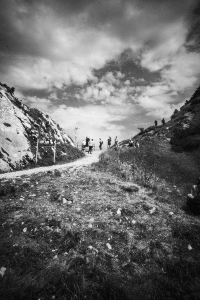 Monochrome Lochkameraaufnahme von Menschen, die auf einem Bergpfad zwischen Felshängen unter einem bewölkten Himmel gehen.