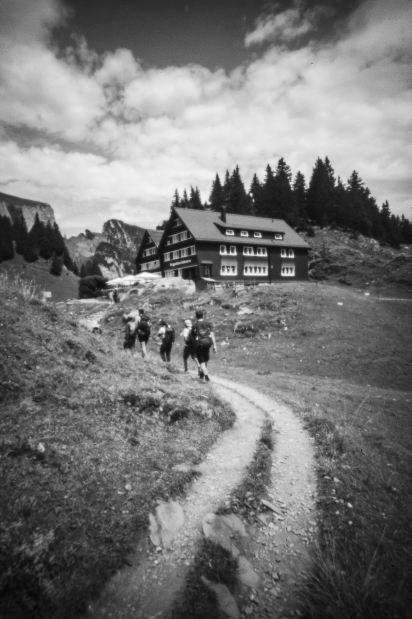 Eine Schwarz-Weiß-Fotografie zeigt eine Gruppe von Wanderern, die auf einem geschwungenen Schotterweg unter einem bewölkten Himmel auf eine große Berghütte zugehen.
