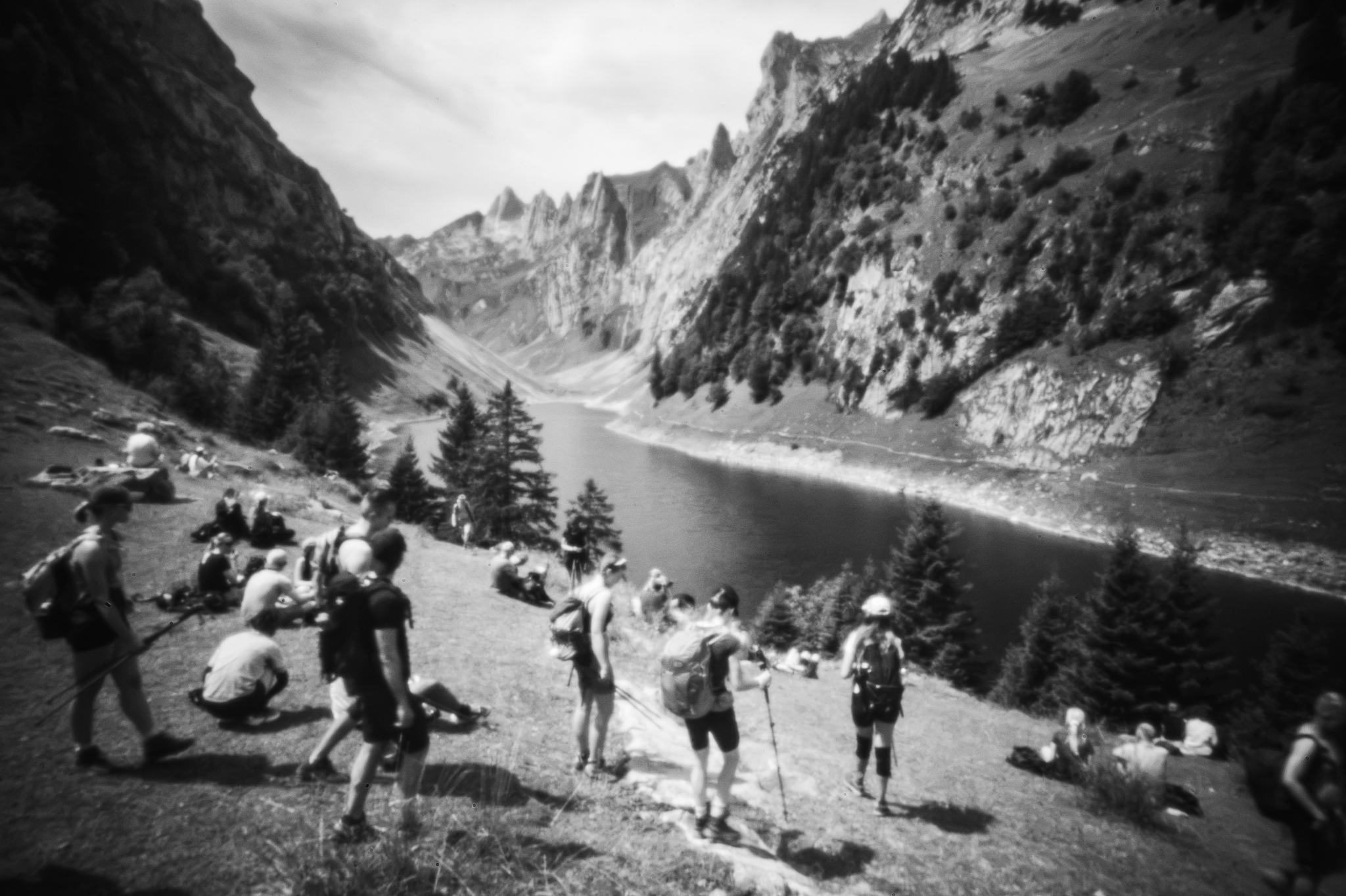 Monochrome, weich gezeichnete Weitwinkelaufnahme von Wanderern, die an der Saxer Lücke auf einem grasbewachsenen Hang neben einem See im Kreis schroffer Gipfel rasten.
