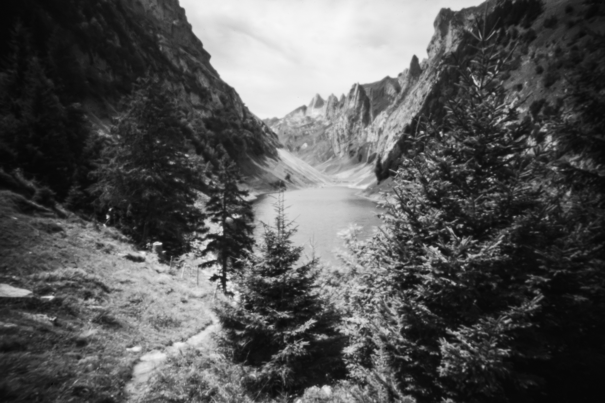 Monochrome Lochkamera-Landschaftsaufnahme eines Bergsees zwischen steilen Felsgipfeln, eingerahmt von Tannen im Vordergrund.