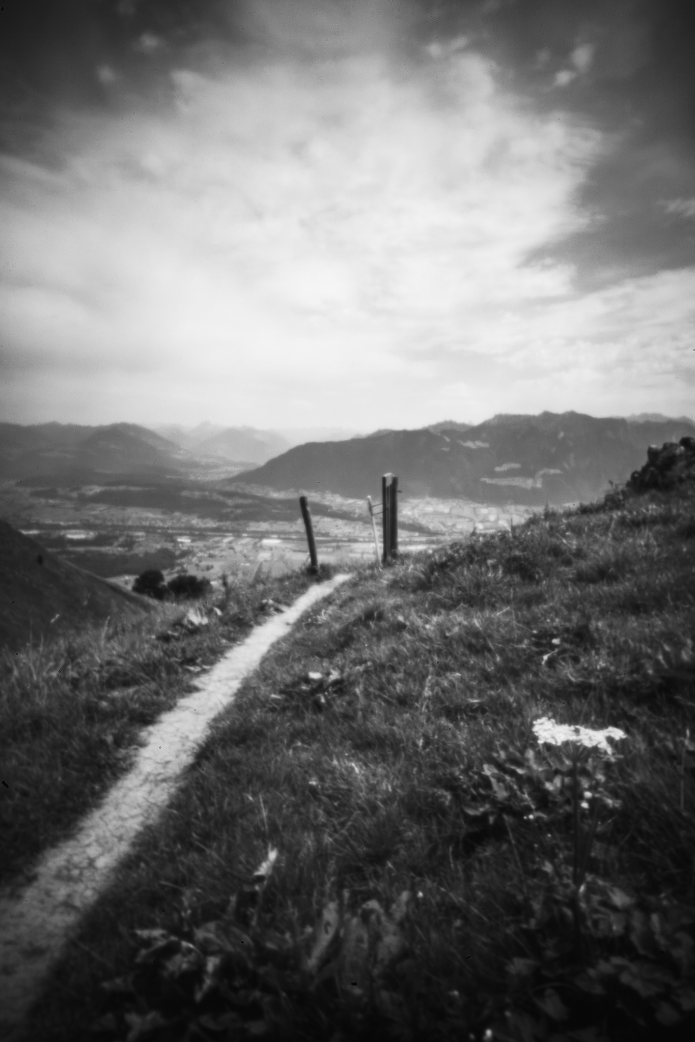Schwarz-Weiß-Landschaft mit einem schmalen Feldweg durch grasbewachsene Hänge, der unter einem bewölkten Himmel zu entfernten Bergen führt, festgehalten im weichen Lochkamera-Stil.