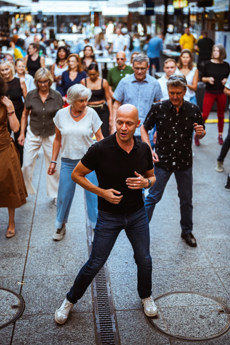 A group of people dancing outdoors on a paved surface, led by a man in a black polo shirt and jeans in the foreground.