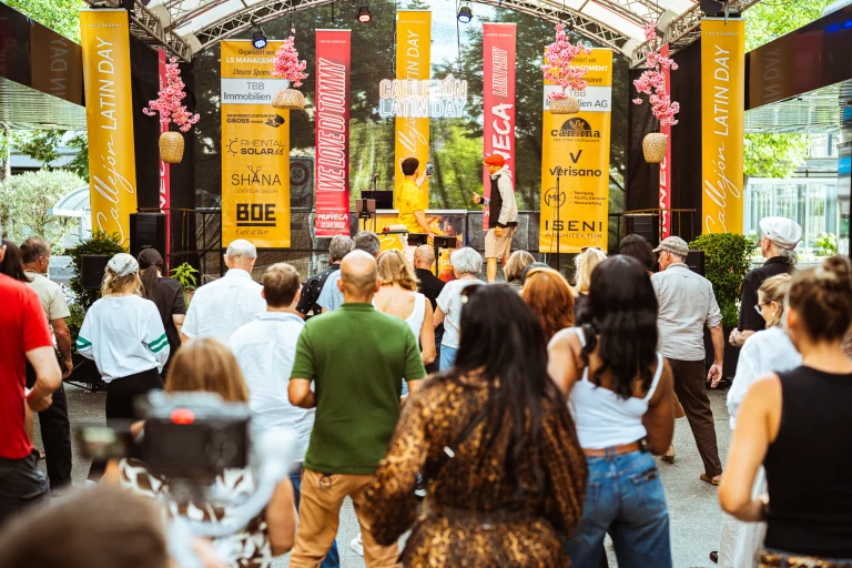 An outdoor audience watches a stage adorned with tall yellow and red banners and neon signage during an outdoor event.