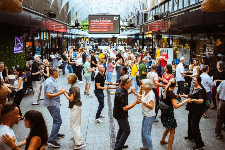 Numerous couples and individuals dance in pairs along a covered outdoor pedestrian shopping arcade.